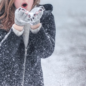 Girl blowing snow off her gloved hands. She's wearing a navy winter coat and scarf and is in a snowy scene