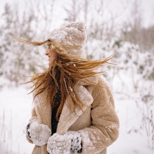 Girl with long blonde hair looking backwards. Dressed in a warm fir lined coat, hat & gloves. In a snowy wintery scene