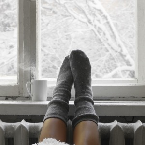 a pair of feet wearing grey wooly socks, resting on top of a radiator in front of a window, showing a snowy wintery scene outside